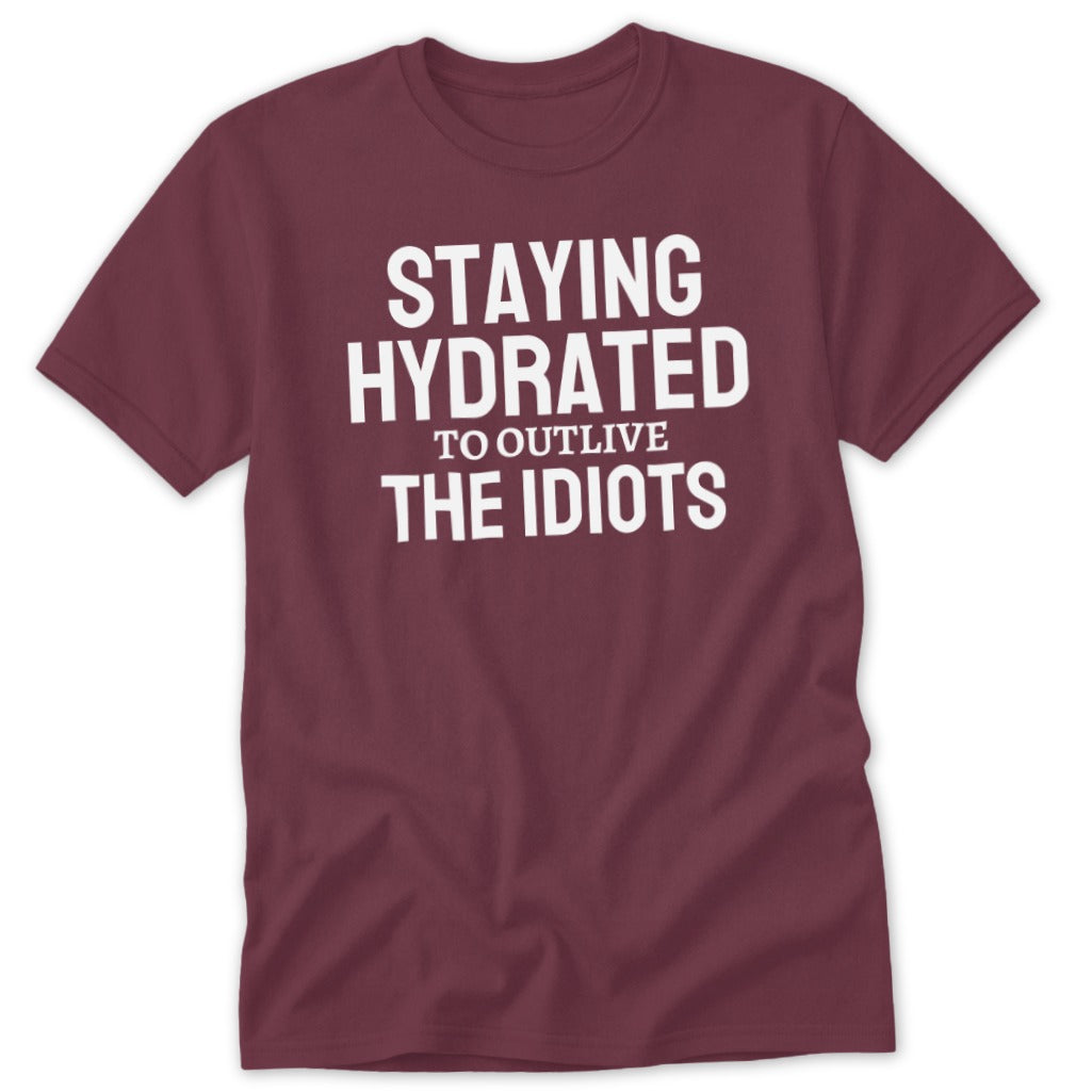 Maroon t-shirt with white text "STAYING HYDRATED TO OUTLIVE THE IDIOTS".