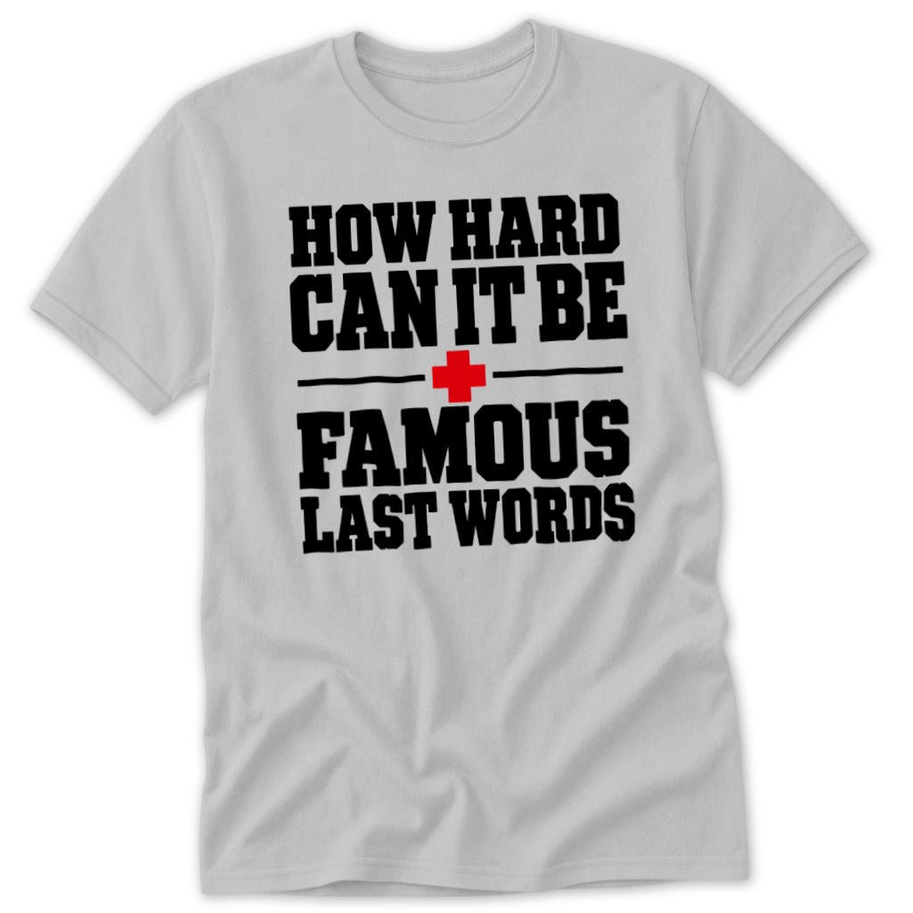 A light gray crewneck t-shirt featuring black block text that reads, HOW HARD CAN IT BE FAMOUS LAST WORDS, separated by a red cross icon.