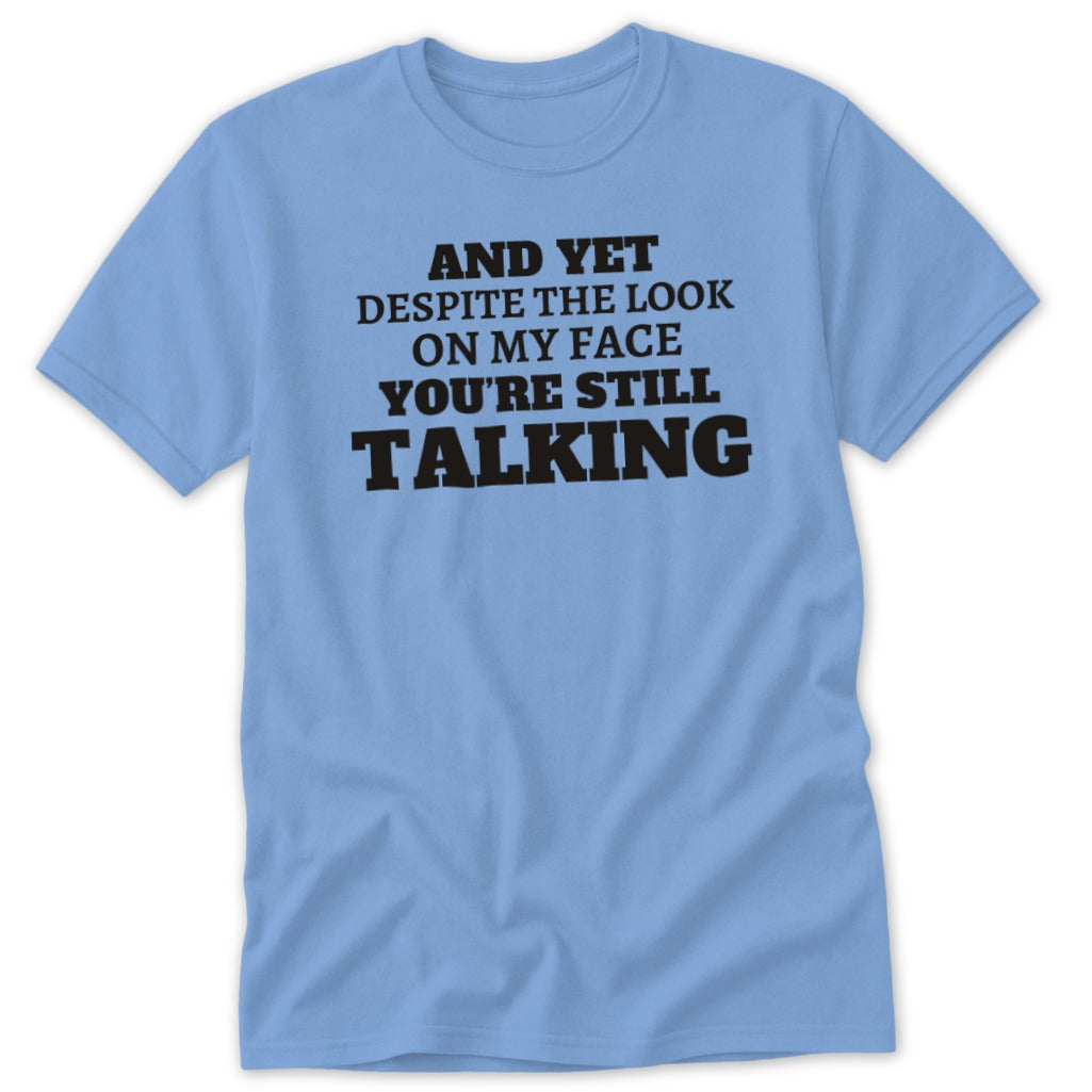 Light blue t-shirt with black text: "And yet despite the look on my face you're still talking"