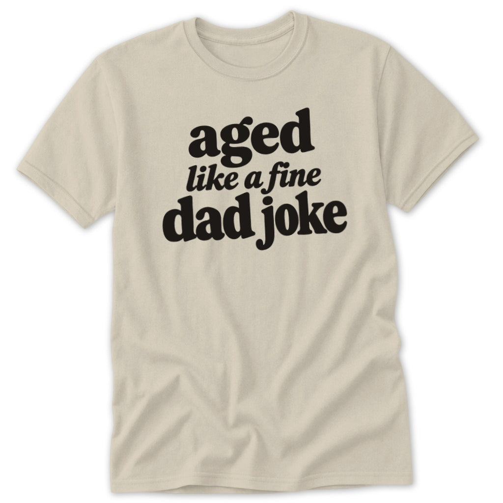 A beige short-sleeve t-shirt with black text on the chest that reads aged like a fine dad joke.