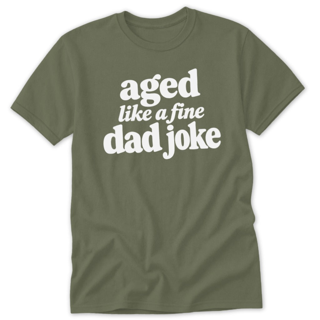 An olive green crewneck t-shirt featuring the white text "aged like a fine dad joke" centered on the chest.